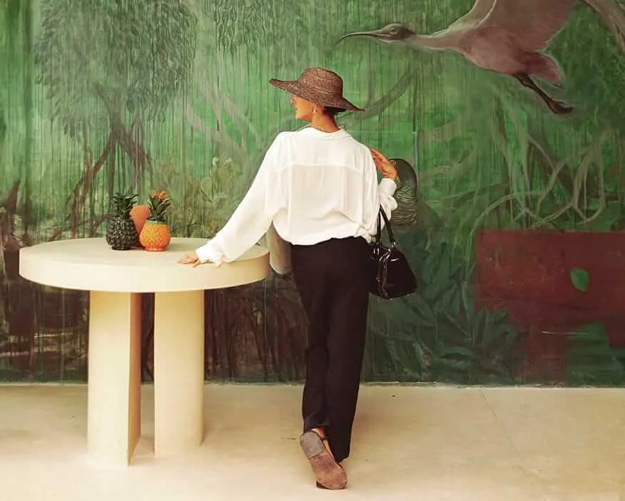 BACAB Tulum – A living example of how well-planned real estate can become a true gem for a city and its community Person in straw hat and white shirt stands at round table with pineapples in front of mural depicting wading birds and wetland vegetation