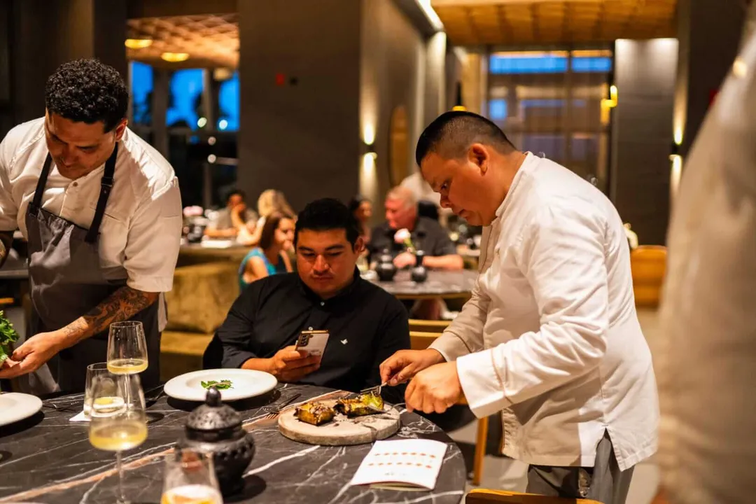 Conrad Tulum Riviera Maya honors Oaxaca through cuisine Two chefs in white chef's coats serve plated dishes to a diner at an upscale restaurant counter during an evening service