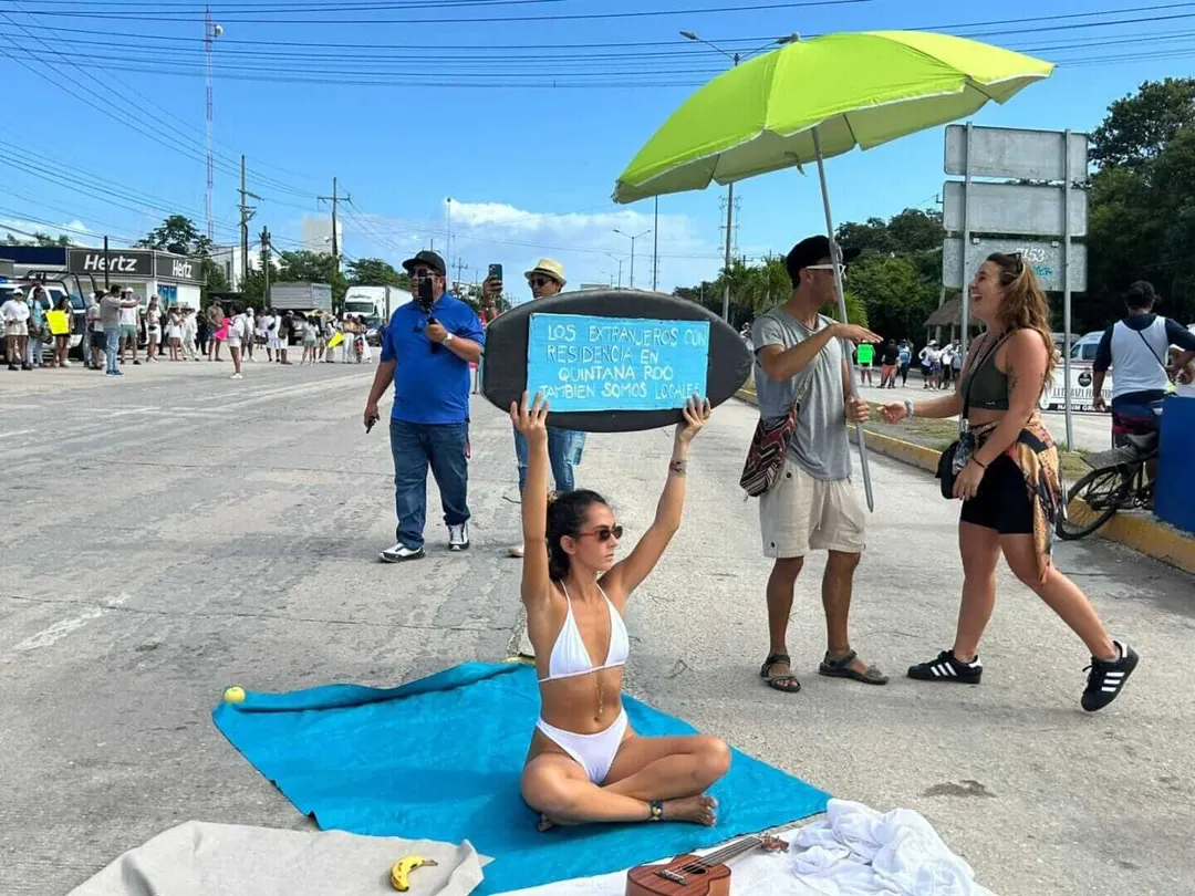 Woman sitting on blue cloth holds a sign while others stand nearby under a yellow umbrella at an outdoor gathering with crowds and vehicles in the background
