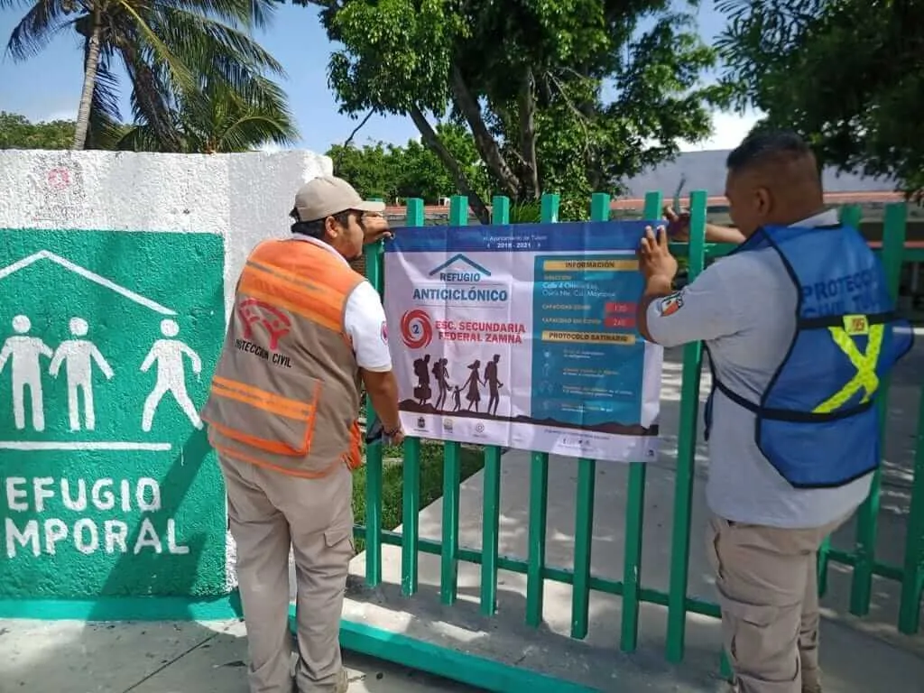 40 Emergency Shelters Stand Ready in Tulum Two workers in safety vests install informational signage on a green fence at what appears to be an emergency shelter facility