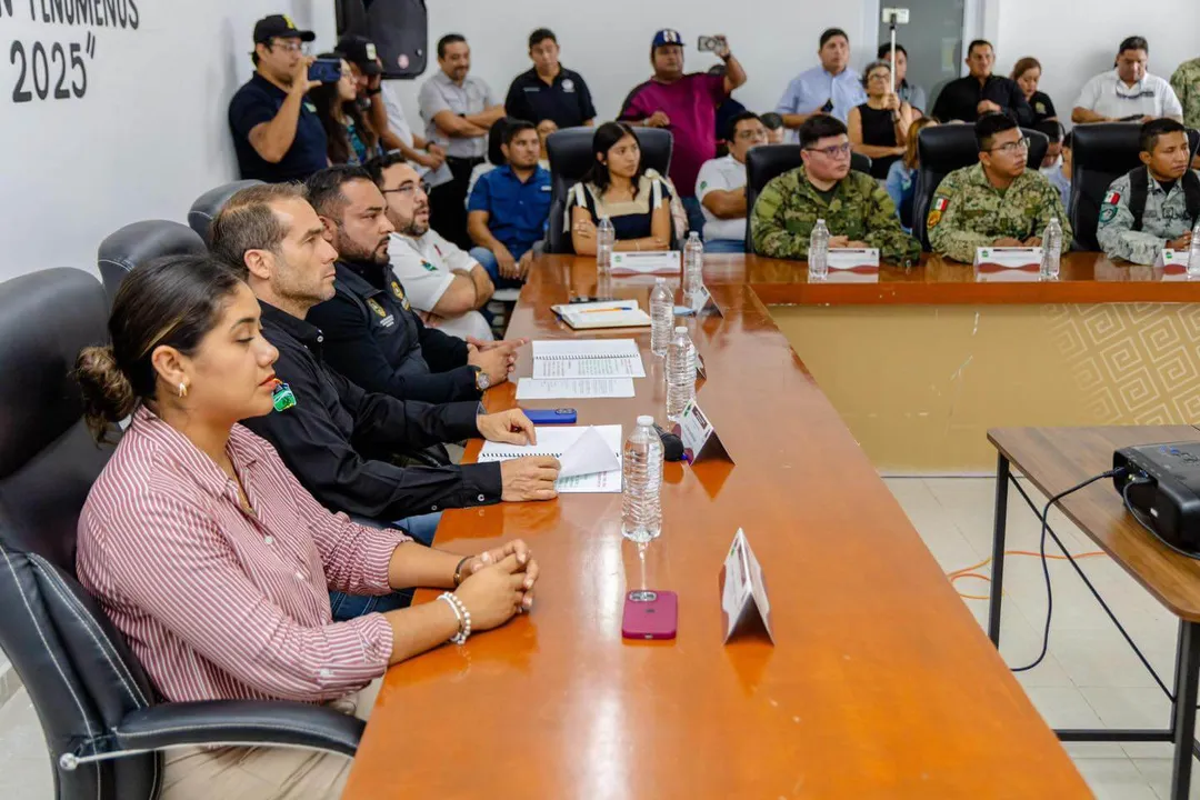 Officials and military personnel sit at a conference table during a hurricane preparedness meeting, with additional attendees standing along the walls of the me