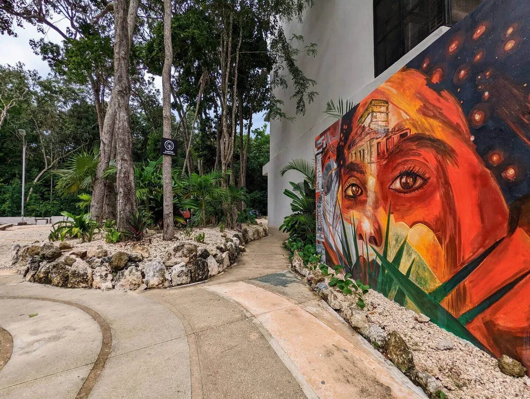 Artists Unite for Climate Change Mural Mural depicting a face in orange and red tones on a white building wall next to a forested pathway with tall trees