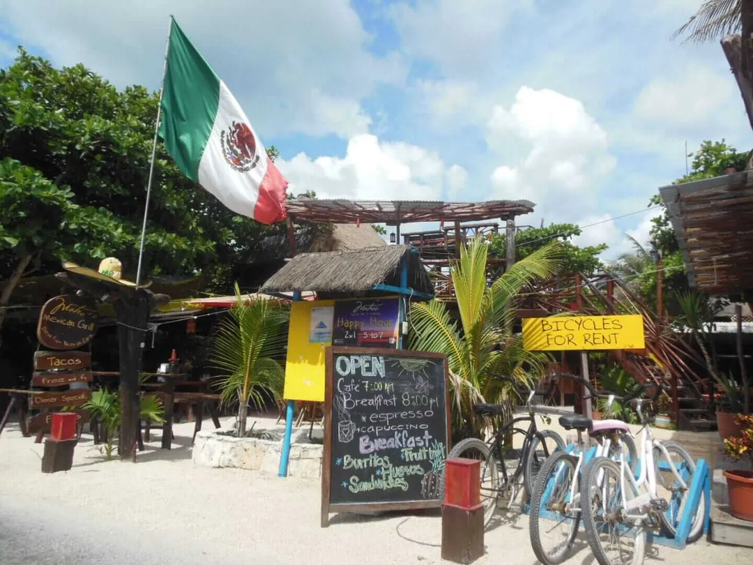 Beach café with Mexican flag flies above rustic wooden structures, bicycles available for rent, and menu board advertising food and beverages