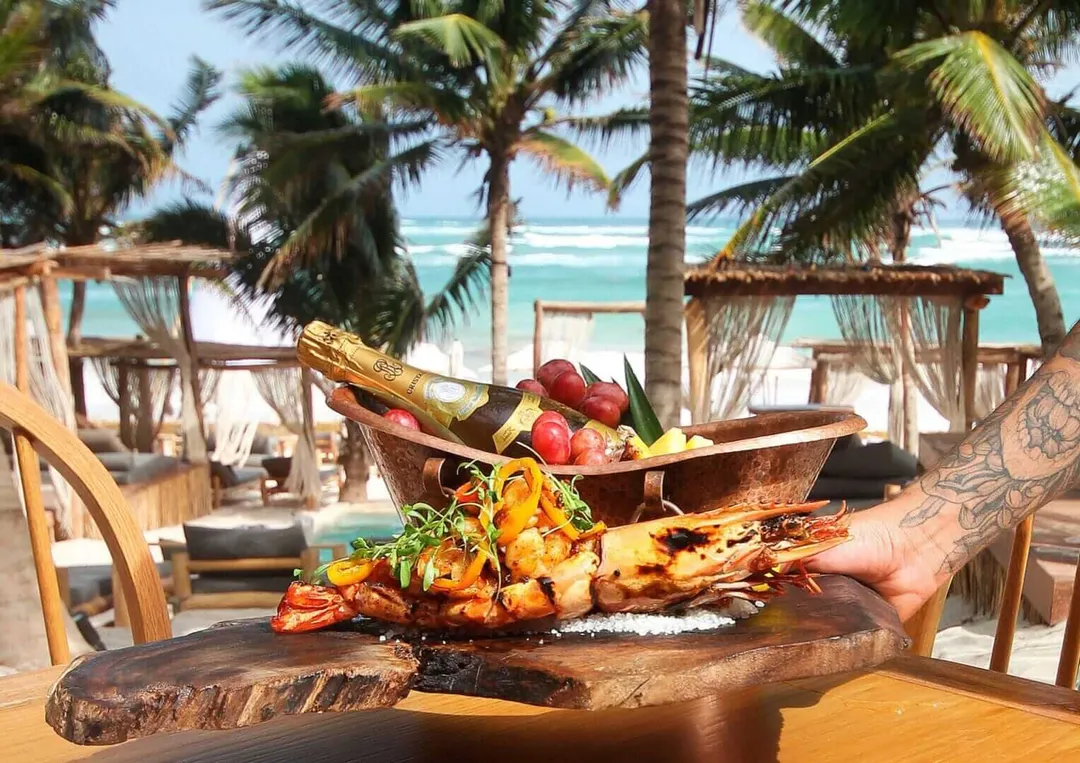The Soul on the Tongue: Tulum Mexican Cuisine in 2025 A tattooed hand holds a wooden plate of grilled seafood and vegetables at a beachfront restaurant with palm trees, tiki structures and turquoise water visible b