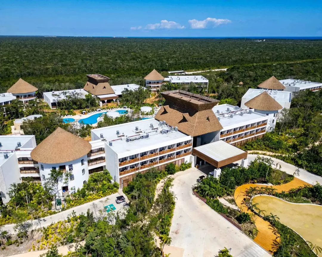 Mundo Maya Brand Relaunch Wows at Tulum Airport Hotel Debut Aerial view of a large resort complex with white buildings featuring thatched roofs, a blue swimming pool, and tropical forest surrounding the property