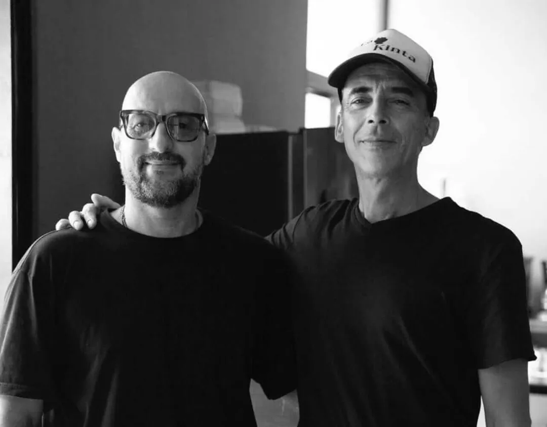 La KINTA Blends German Roots and Mayan Soul with Chef René Piepenburg Two men stand together indoors with their arms around each other's shoulders, one wearing glasses and the other wearing a cap, both in black shirts