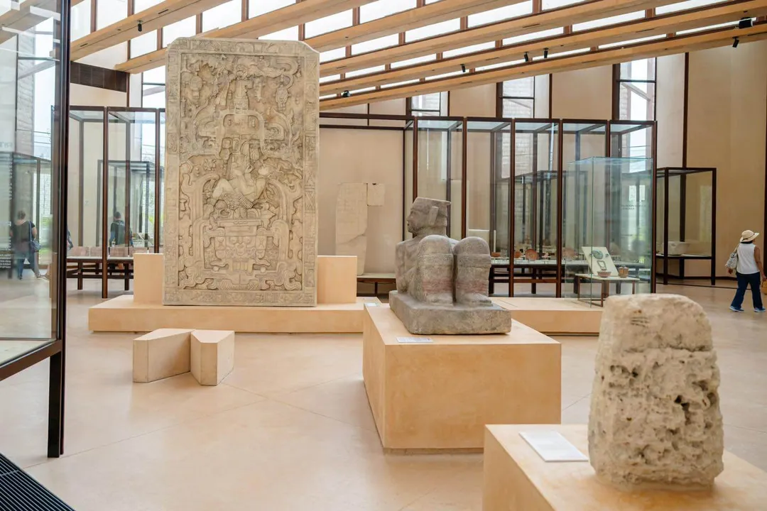 Museum gallery with carved stone artifacts and fragments displayed on tan pedestals beneath wooden beams and skylights