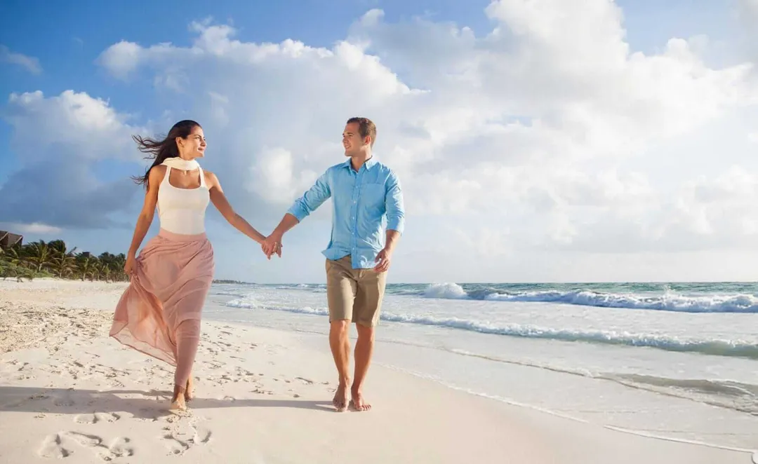 A couple holds hands while walking along a sandy beach with ocean waves and cloudy sky in the background