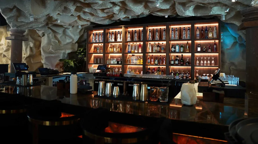 New Tulum‑Inspired Restaurant and Bar Tago Opens in Houston A bar interior features an illuminated shelving unit displaying bottles, with a dark counter in the foreground and textured stone walls above