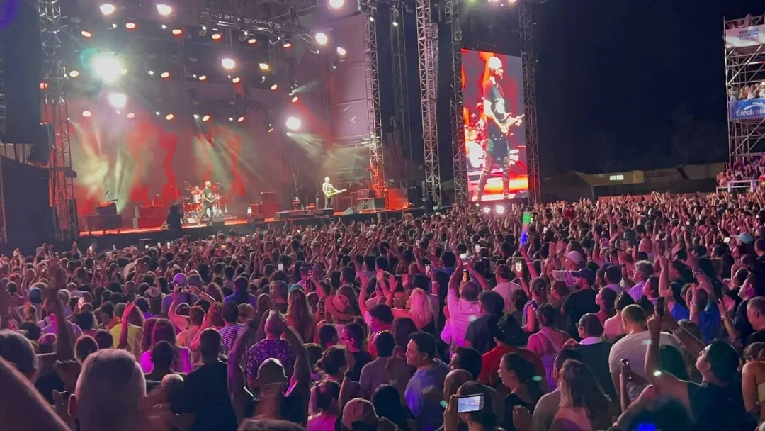 Large crowd of concertgoers with raised hands and phones watches performers on an illuminated stage at a nighttime outdoor music festival