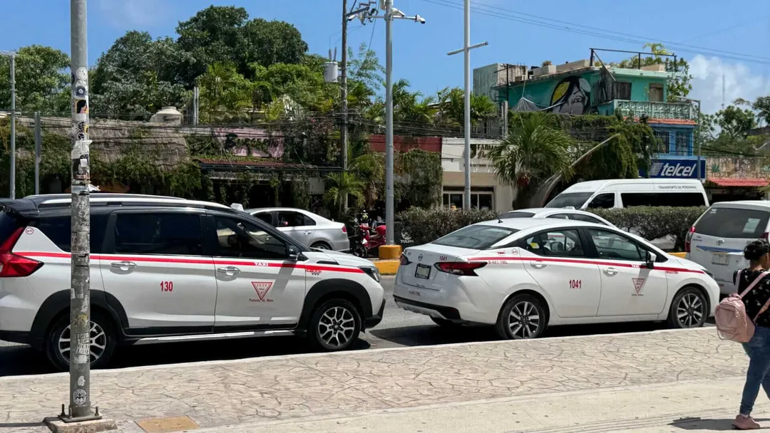 Tulum Taxi Drivers Face Economic Dip Before Summer Boost Two white taxis with red stripes parked on a paved street with buildings and vegetation visible in the background