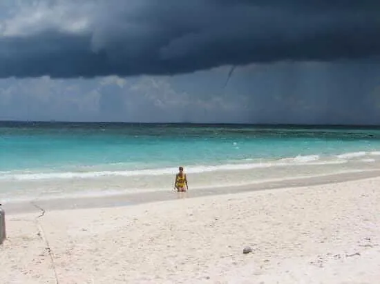 Person wading in turquoise water on a sandy beach under dark storm clouds approaching over the ocean