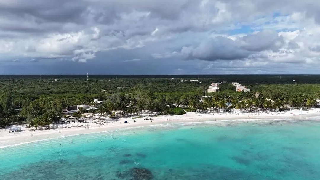 Turquoise water and white sand beach lined with palm trees and beachfront buildings under cloudy sky