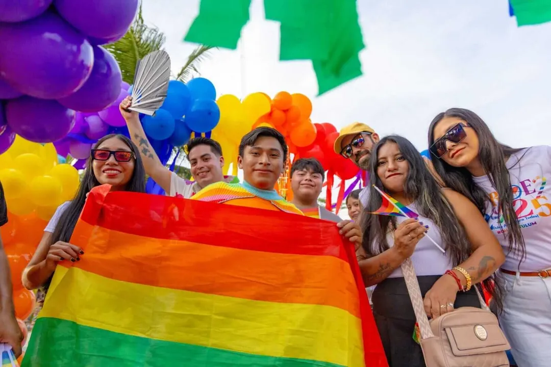 Tulum Pride Parade Unites Locals Tourists and Activists in Vibrant Celebration Group of people holding a rainbow flag at an outdoor event with colorful balloons and decorations overhead