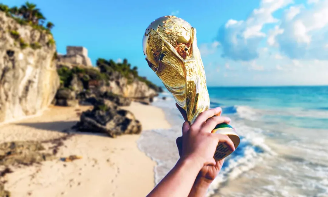 A person holds a gold trophy on a beach with rocky cliffs, ocean waves and palm trees visible in the background