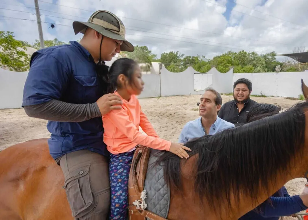 How Equine‑Assisted Therapy Is Empowering Children and Adults in Tulum An instructor guides a young rider on a brown horse while two men watch during an equine therapy session in a sandy arena