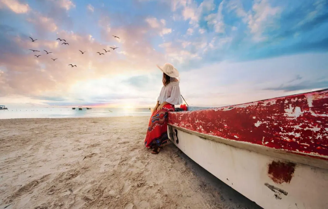 Tulum Was Never a Destination; It Was a Mirror Person in white shirt and hat stands beside a weathered red and white boat on a sandy beach at sunset, with birds flying overhead