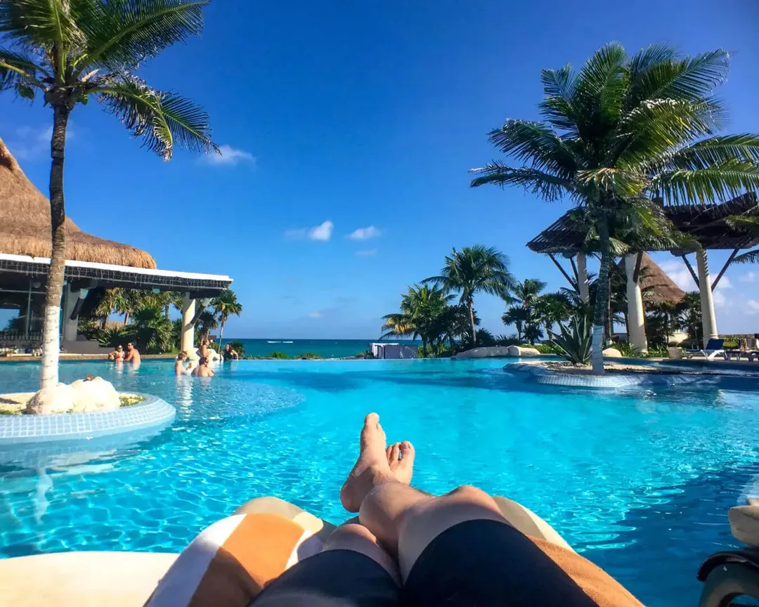 Why travelers are choosing Tulum all inclusive resorts this summer Person relaxing by a large resort pool with palm trees, thatched-roof structures, and ocean visible in the background under clear blue sky