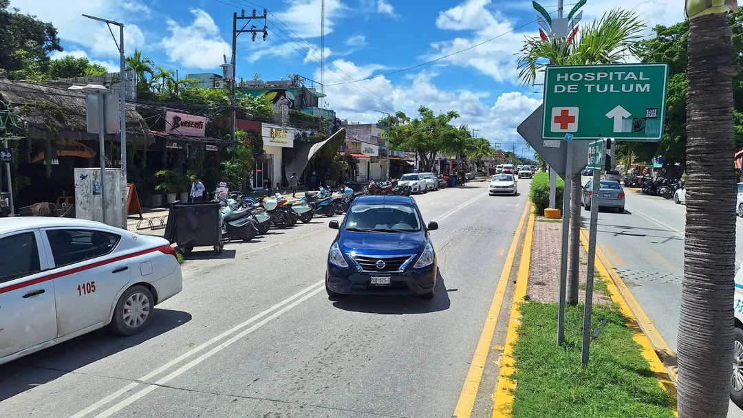 A street scene with a blue sedan and white emergency vehicle driving past a green hospital directional sign, with palm trees and storefronts visible along the r
