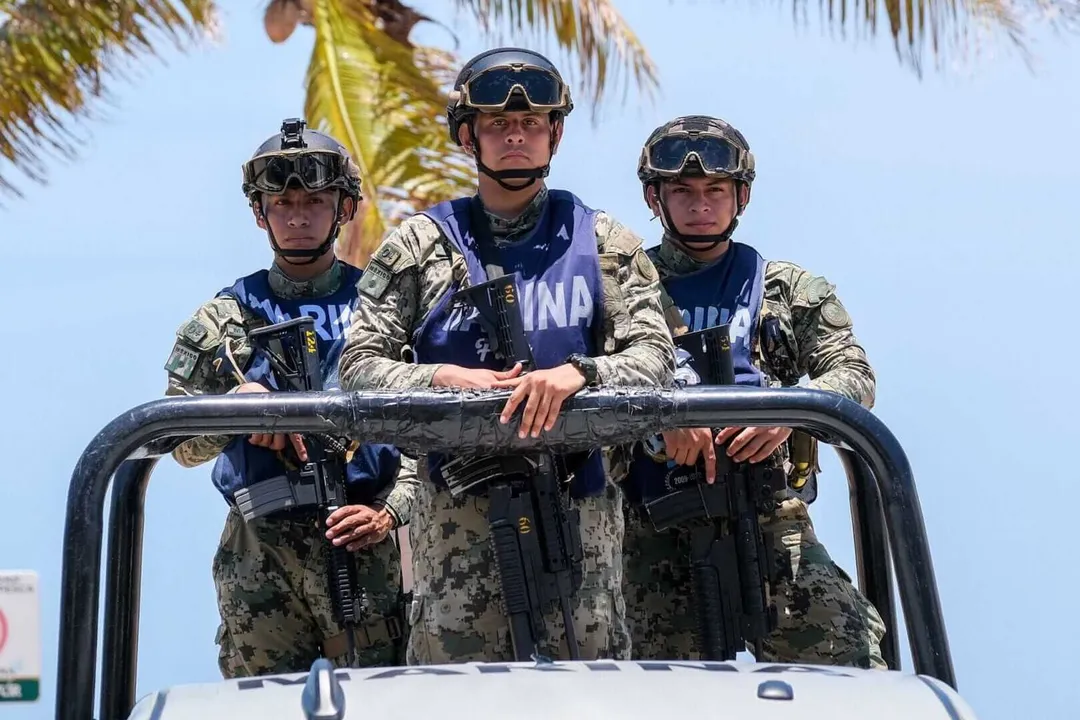 Three armed security personnel in camouflage uniforms and tactical gear stand in a vehicle with palm trees visible behind them