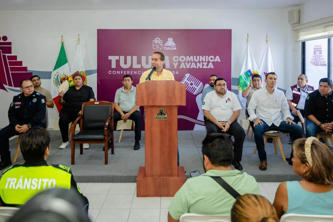 A man speaks from a wooden podium during a press conference while seated officials and attendees listen in a room with a purple banner reading "Tulu Comunica y