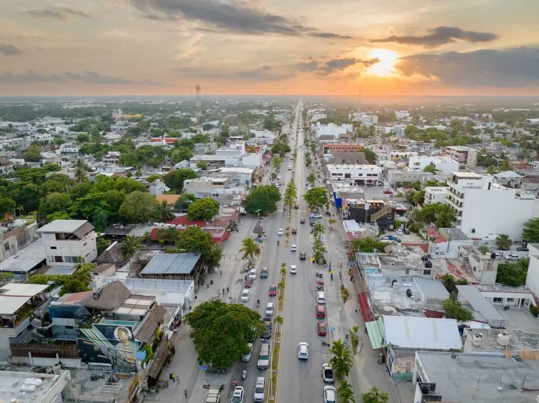 Aerial view of a wide street lined with trees and vehicles running through a sprawling town at sunset, with buildings on either side and green landscape extendi