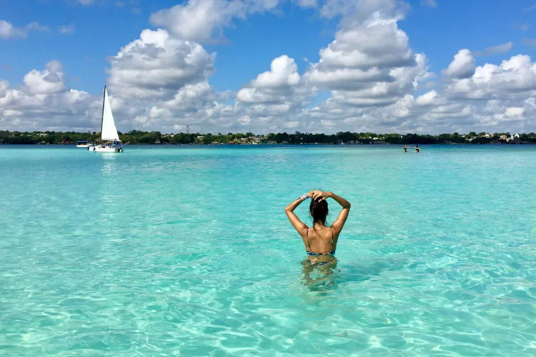 Person wading in turquoise water with sailboat and waterfront homes visible in the distance under cloudy sky