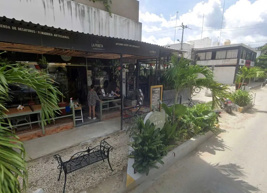 A small storefront with a black awning sits along a street lined with potted plants, with a metal bench in front and buildings visible in the background under a