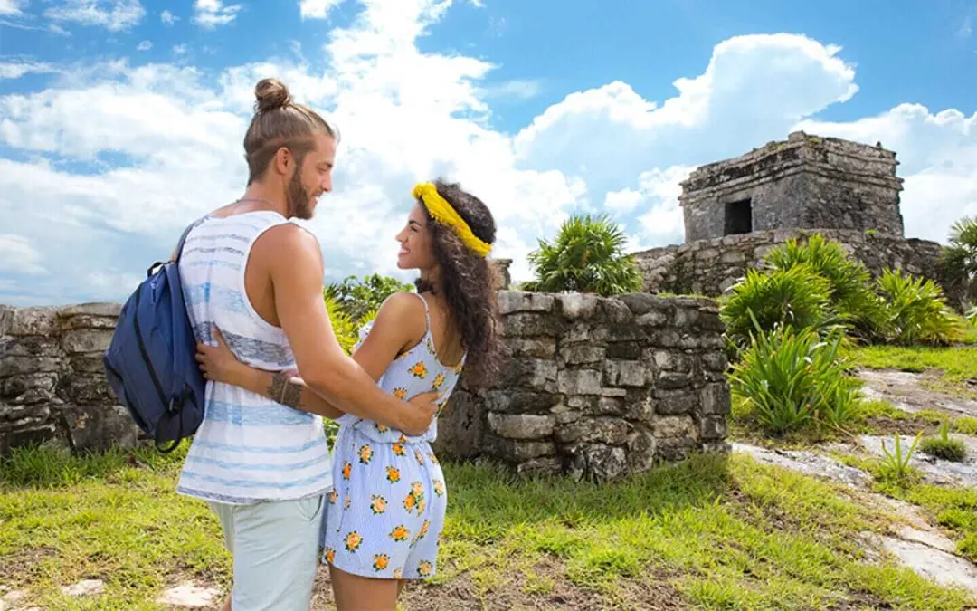 Two tourists stand facing each other on grassy ground near ancient stone ruins with a tower structure visible in the background