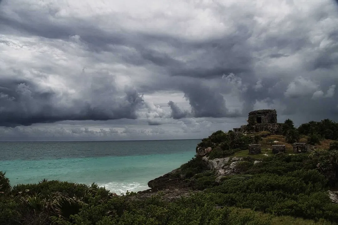 Tulum is sailing through the biggest storm in decades without a clear destination Ruins of a stone structure sit atop a rocky coastal cliff overlooking turquoise water beneath dark storm clouds