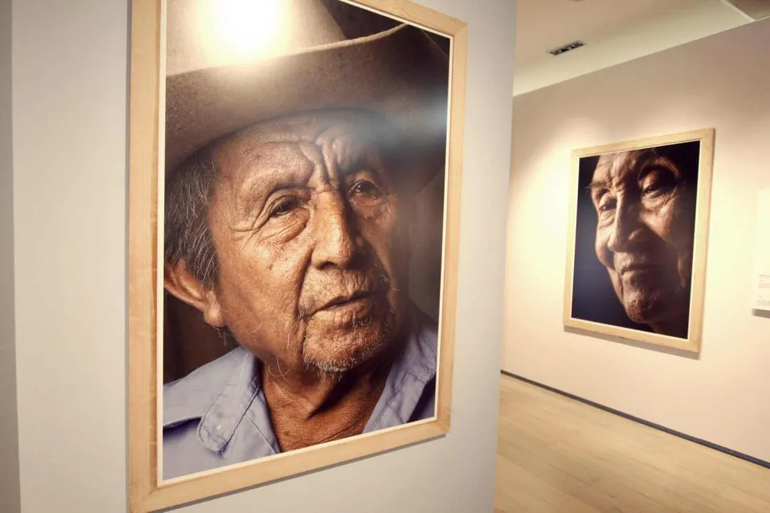 Large portrait photograph of an older man with lined face and gray hair wearing a light blue shirt, displayed in a wooden frame on a gallery wall with a smaller