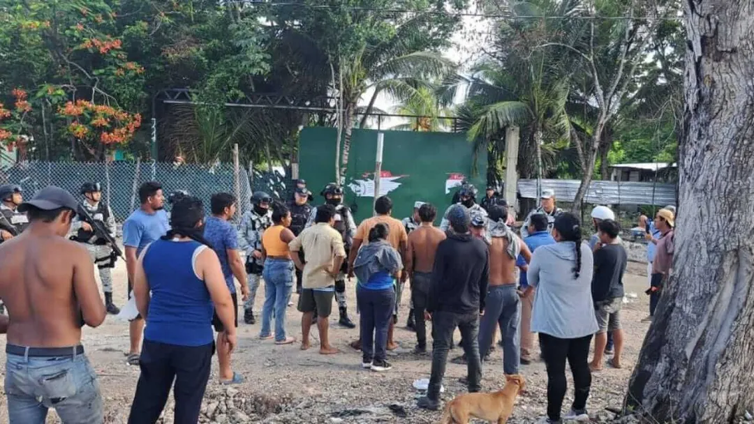 A crowd of people gathers in a dirt lot surrounded by trees and green buildings during what appears to be a community assembly or dispute