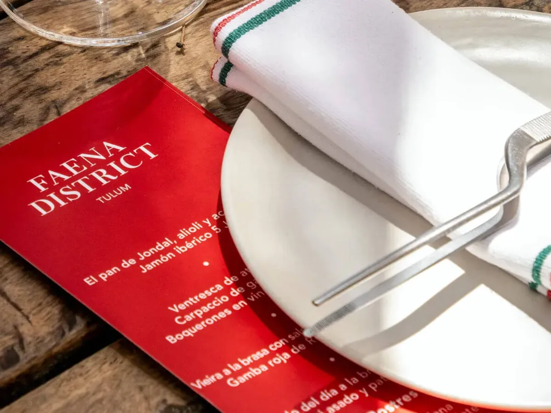 Faena Tulum Launches in Ibiza and Sets a New Standard for Luxury Real Estate in Tulum A red brochure for Faena District Tulum lies on a wooden surface next to folded white kitchen towels with striped trim and a white plate with fork
