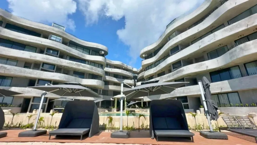 Modern apartment complex with curved balconies surrounding a courtyard with lounge chairs and umbrellas under blue sky