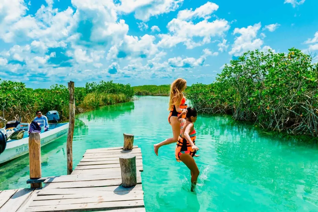 Woman running off a wooden dock into turquoise water surrounded by mangrove trees, with a boat moored nearby under blue sky