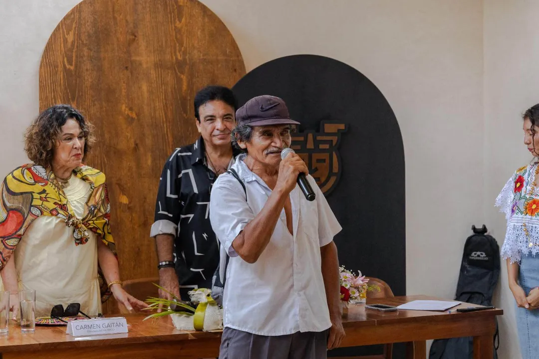 The Man Who Carried a War in His Silence Man wearing white shirt and purple cap speaks into microphone at wooden table during panel event with three other people standing beside him