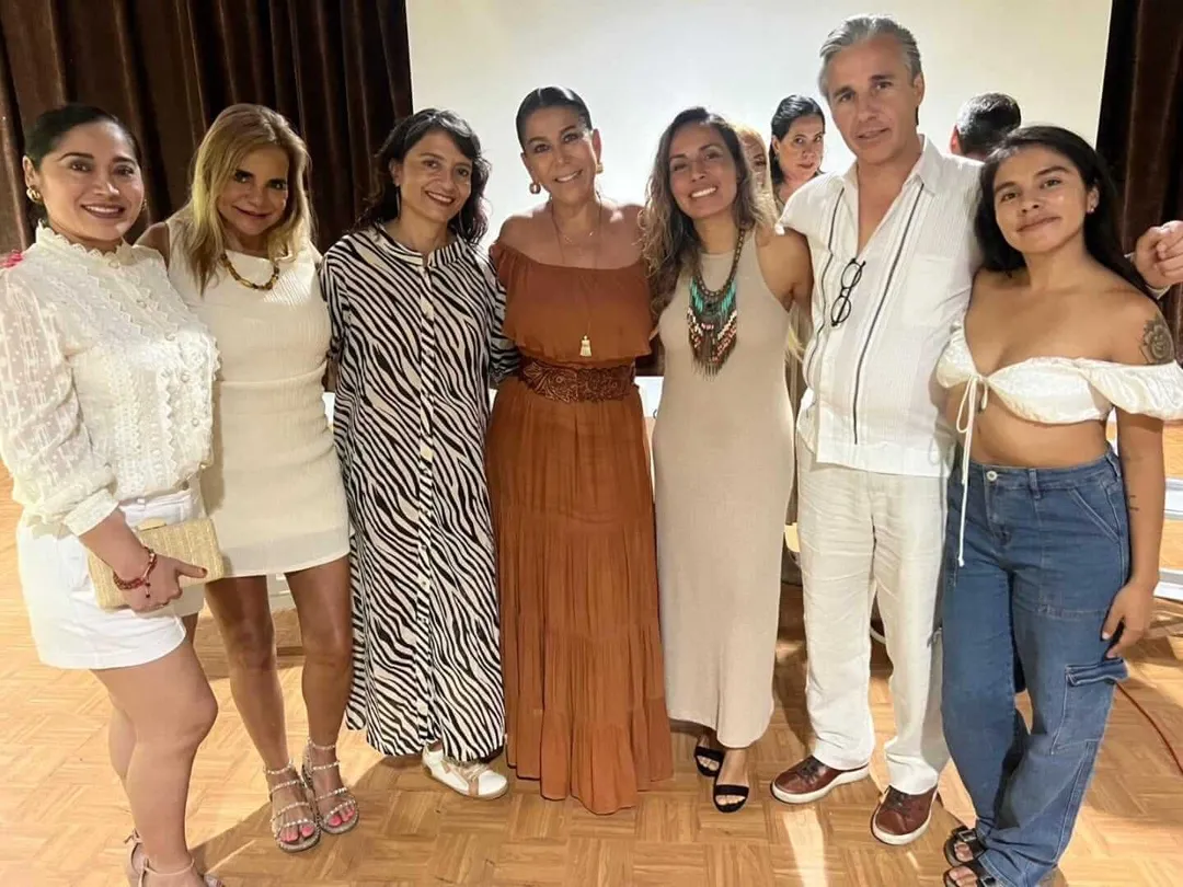 New Alliances Help Tulum Navigate Shifting Tourism Tides Eight people stand together indoors smiling at the camera in front of a white wall and brown curtains