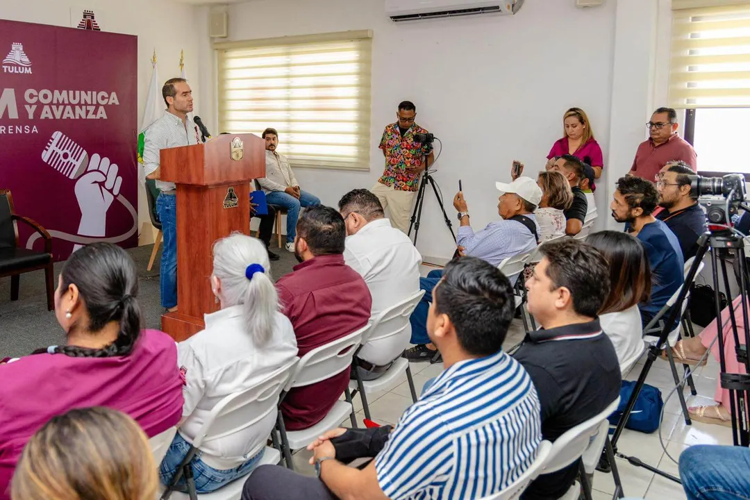 Tulum Balances on the Edge of Chaos With Sand in Its Shoes A man speaks at a wooden podium during a public meeting as seated attendees listen, with camera operators and standing observers present in an indoor venue