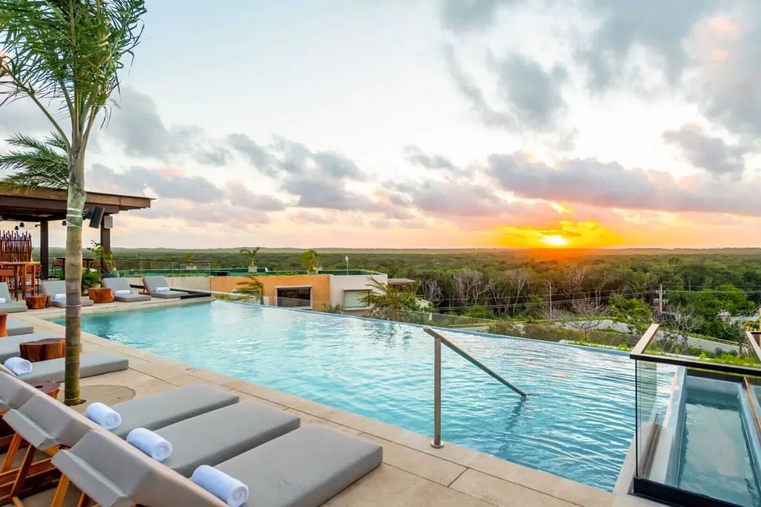 Rooftop pool with lounge chairs overlooks a sprawling landscape at sunset with scattered clouds above