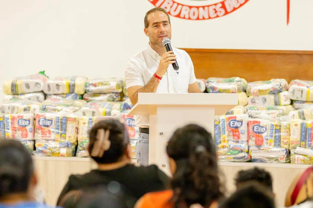 The Hands That Keep Tulum Alive Receive a Small Relief Man speaking at a podium with a microphone in front of stacked food packages, addressing an audience from behind