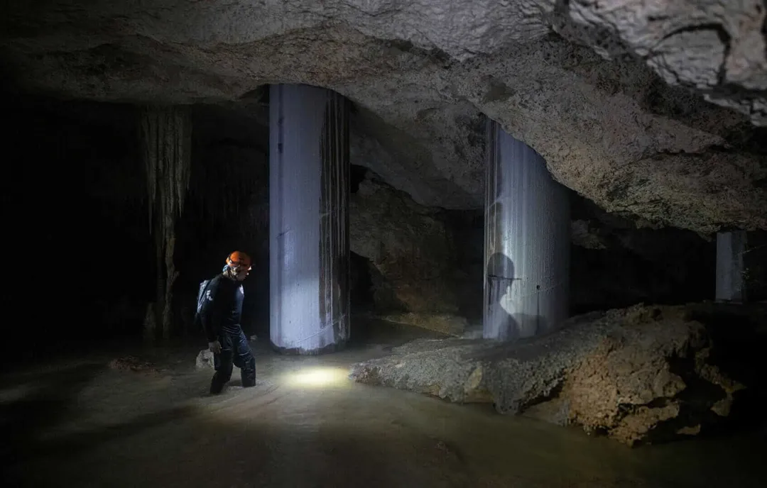 Beneath the Tren Maya Tracks Lies a World on the Edge of Collapse Person with headlamp exploring an underground cave chamber with cylindrical concrete support pillars and rocky debris