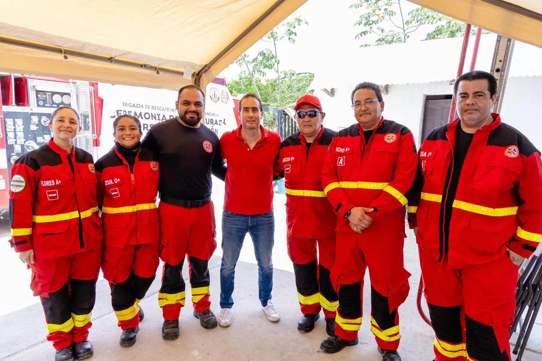 Seven firefighters in red and yellow uniforms stand together under a tent with one man in casual clothing in the center
