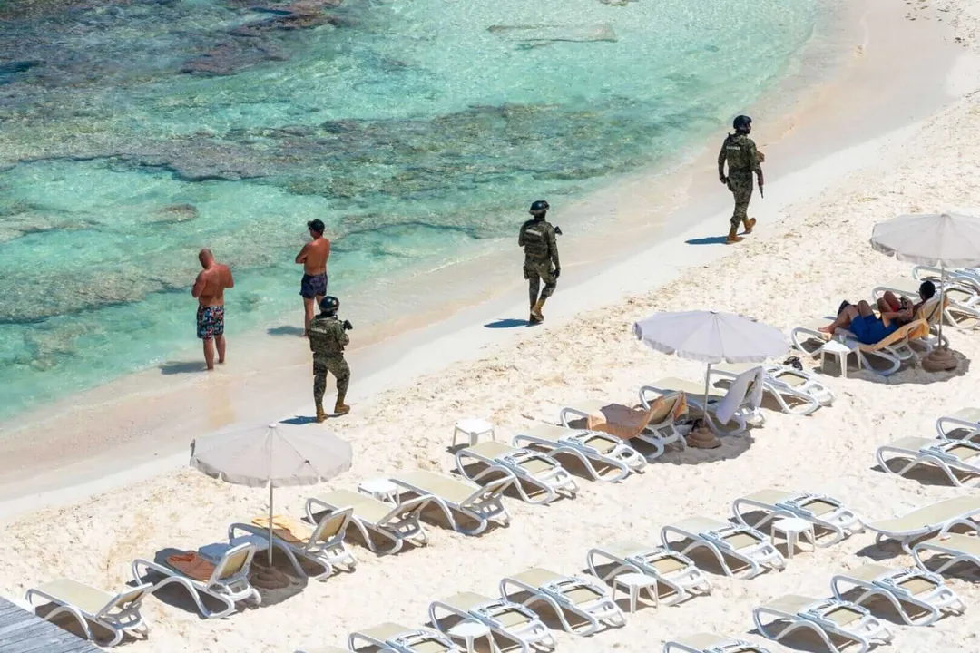 Uniformed personnel walk along a sandy beach lined with lounge chairs and umbrellas as visitors wade in shallow turquoise water