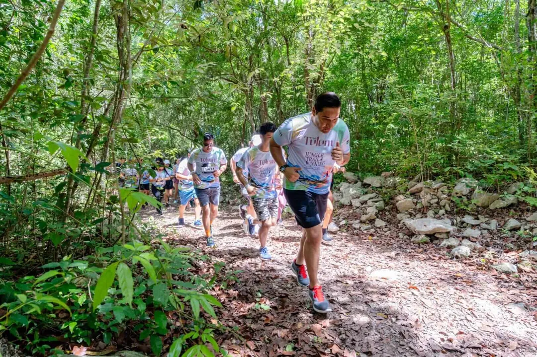A group of trail runners jog along a rocky forest path surrounded by dense green vegetation and trees