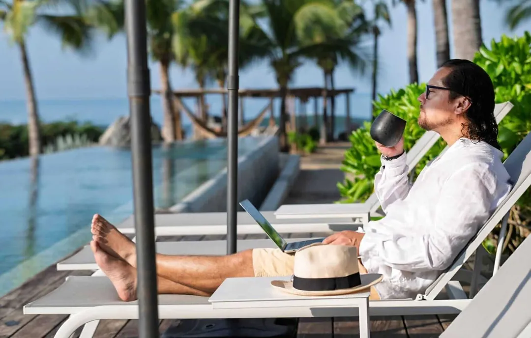 Man in white shirt and sunglasses reclines on a lounge chair holding a laptop while overlooking a beachfront pool area with palm trees and ocean views