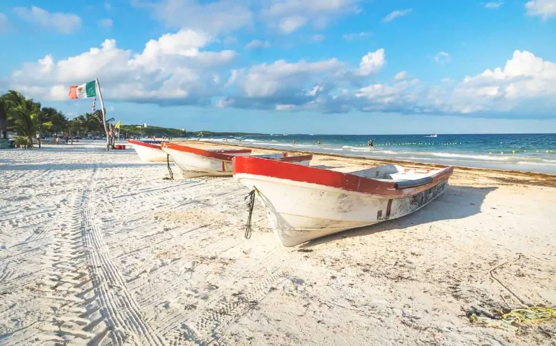 Tulum Beaches Earn White Flag Certification for Conservation Efforts Fishing boats with red and white hulls sit on sandy beach with calm turquoise water and palm trees in the distance under blue sky with white clouds