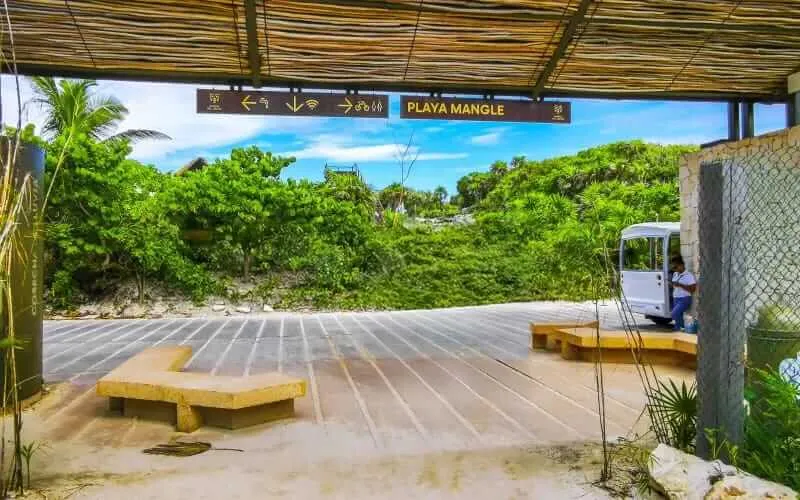Tulum Offers Free Pedestrian Access to Mangle Beach Amid Protest Plans A wooden platform and seating area sit on sand under a thatched roof shelter, with a sign reading "Playa Mangle," dense tropical vegetation visible beyond