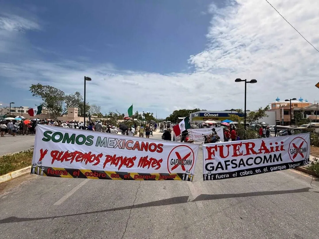 Protesters hold large banners with Spanish text in a street gathering under a partly cloudy sky, with buildings and street lights visible in the background