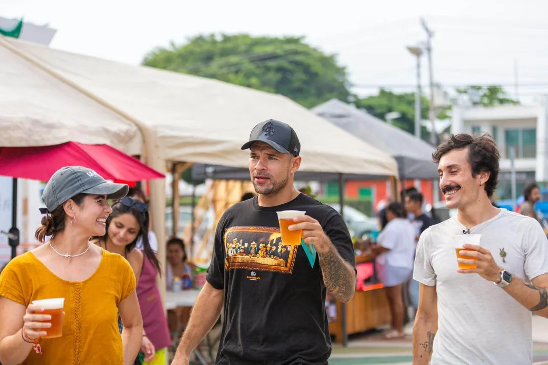 Craft Beer with Maya Roots Brings Buzz Back to Tulum’s Beekeeping Tradition Three people walk through an outdoor festival holding drinks, with market tents and crowds visible in the background