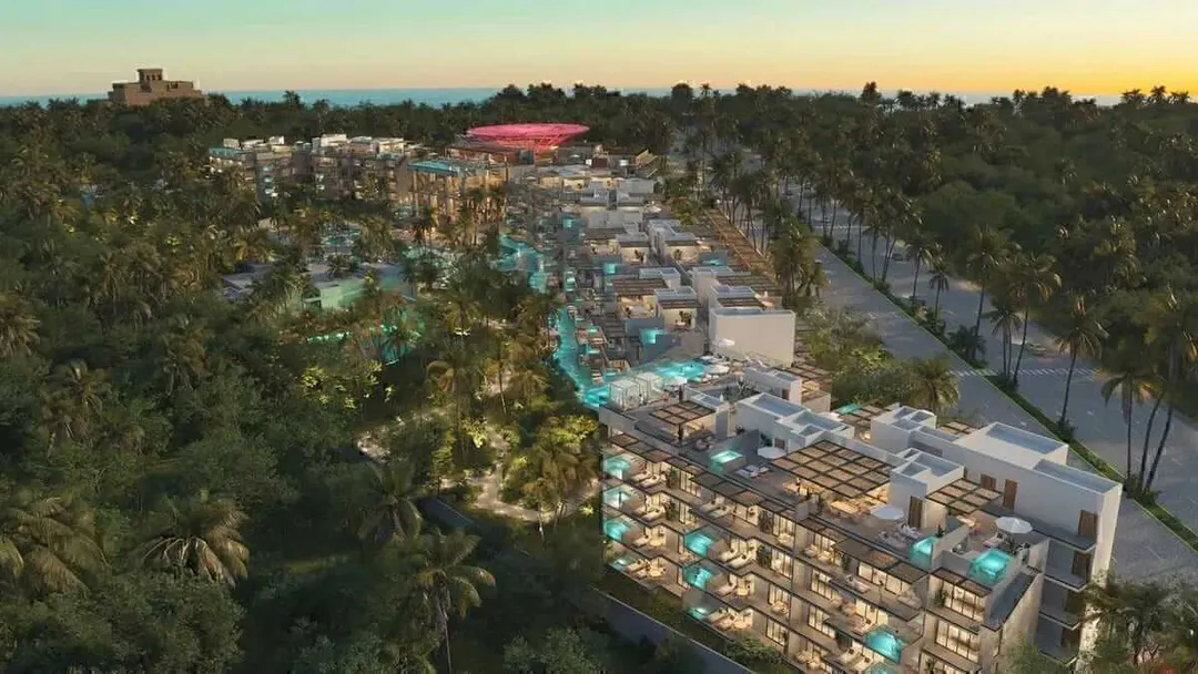 Aerial view of a large resort development with multiple buildings, pools, and palm trees at dusk, surrounded by forested landscape